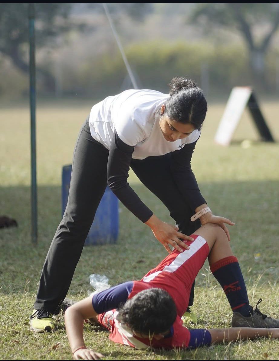Dr. Vedika Gujarathi performing a pitch-side hamstring stretch on an athlete lying on the grass.